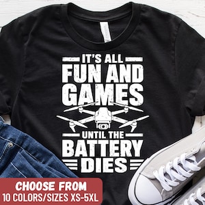 May include: A black t-shirt featuring a white graphic of a quadcopter drone centered between the text "IT'S ALL FUN AND GAMES" at the top and "UNTIL THE BATTERY DIES" at the bottom, with a distressed, weathered font style for the lettering.