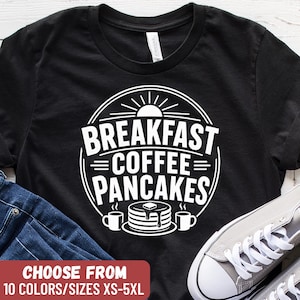 Può includere: T-shirt nero con grafica bianca circolare con la scritta "BREAKFAST COFFEE PANCAKES". Il design include una pila di pancake, tazze di caffè e un sole. Il testo "CHOOSE FROM 10 COLORS/SIZES XS-5XL" è in basso.