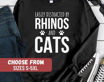Può includere: Felpa nera con testo bianco che recita "Easily Distracted by Rhinos and Cats" con impronte di zampe. Il testo "Choose From Sizes S-5XL" è su un rettangolo rosso.