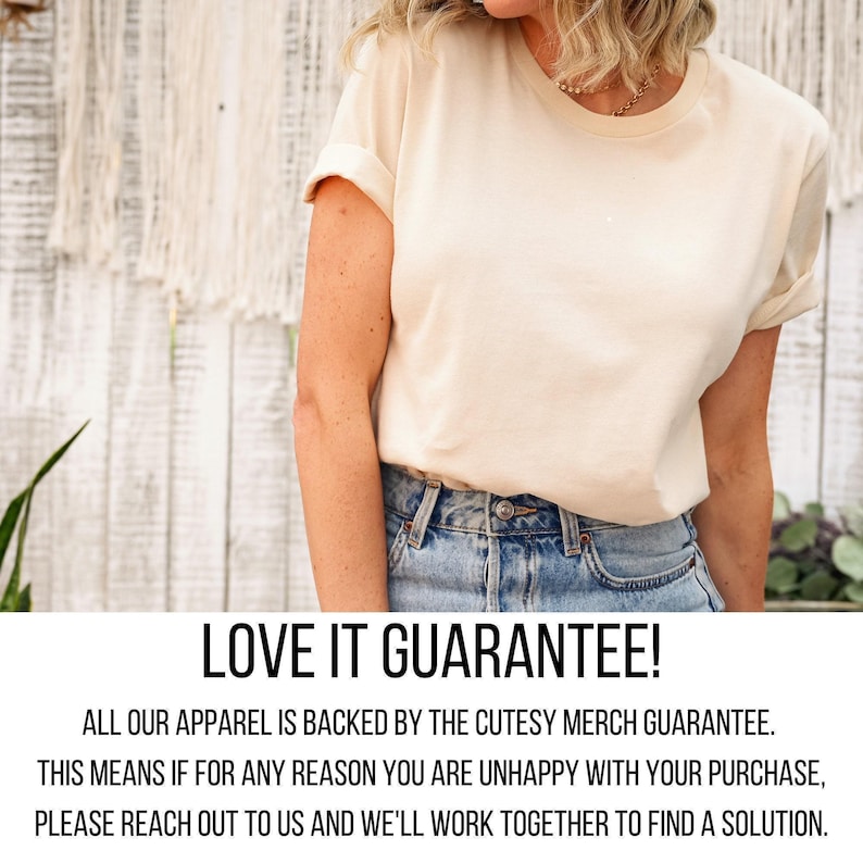 A woman wearing a white t-shirt and blue jeans stands in front of a white wall with a plant.