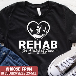 May include: Black t-shirt with a white graphic of a heart, heartbeat, and figure, with the word "REHAB" and the phrase "It's A Work Of Heart." The shirt also has the text "CHOOSE FROM 10 COLORS/SIZES XS-5XL."