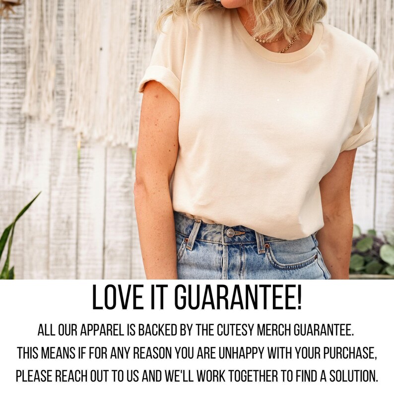 A woman wearing a white t-shirt and blue jeans stands in front of a white wall with a plant.