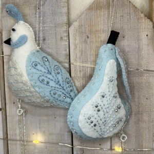 May include: Two felt ornaments, a blue and white bird and a blue and white pear, hanging from a string of lights against a wooden background.