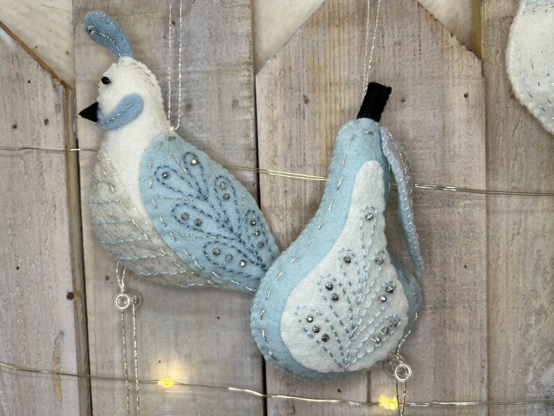 May include: Two felt ornaments, a blue and white bird and a blue and white pear, hanging from a string of lights against a wooden background.