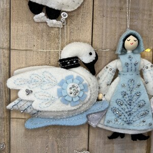 May include: A set of felt Christmas decorations featuring a white swan with blue accents, a blue and white dressed doll holding a bucket, and a blue and white circular decoration with a snowflake design.