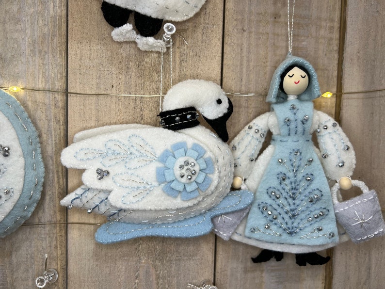 May include: A set of felt Christmas decorations featuring a white swan with blue accents, a blue and white dressed doll holding a bucket, and a blue and white circular decoration with a snowflake design.