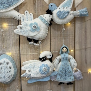 May include: A set of five felt ornaments in a winter theme. The ornaments include a swan, a bird, a girl, and two eggs. The ornaments are all white with blue accents and are decorated with beads and sequins.