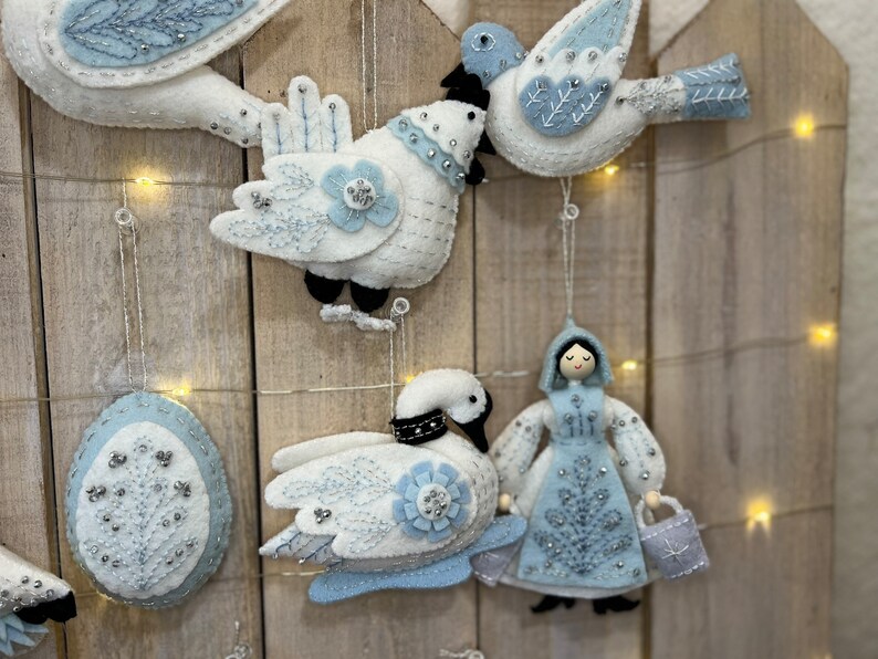 May include: A set of five felt ornaments in a winter theme. The ornaments include a swan, a bird, a girl, and two eggs. The ornaments are all white with blue accents and are decorated with beads and sequins.