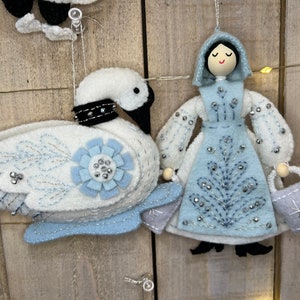 May include: A felt ornament set featuring a white swan with blue and silver accents and a blue felt doll with a white felt dress and a silver bag. The doll has a painted face with black hair and a black hooded cloak.