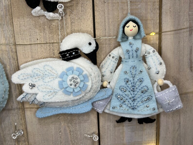 May include: A felt ornament set featuring a white swan with blue and silver accents and a blue felt doll with a white felt dress and a silver bag. The doll has a painted face with black hair and a black hooded cloak.