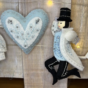 May include: A set of three felt Christmas decorations, including a blue heart with white embroidery, a white figure with a black top hat, and a white tree with blue leaves.