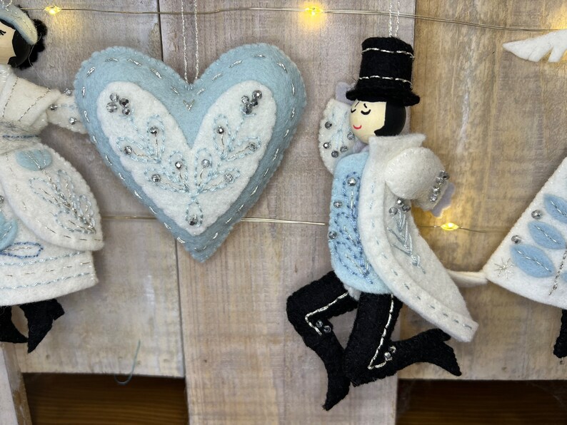 May include: A set of three felt Christmas decorations, including a blue heart with white embroidery, a white figure with a black top hat, and a white tree with blue leaves.