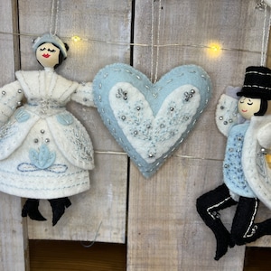 May include: A set of three felt ornaments, including a blue heart with white embroidery, a white felt woman with a blue dress and a black hat, and a white felt man with a blue suit and a black top hat.