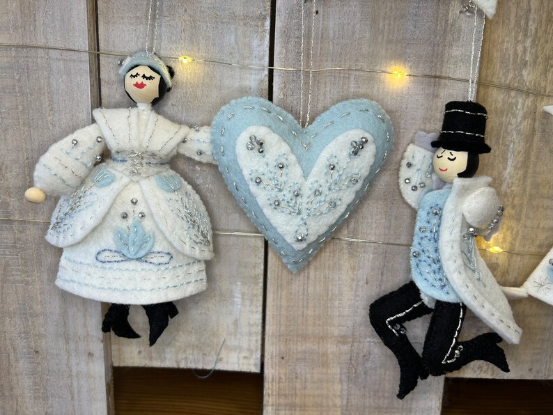 May include: A set of three felt ornaments, including a blue heart with white embroidery, a white felt woman with a blue dress and a black hat, and a white felt man with a blue suit and a black top hat.