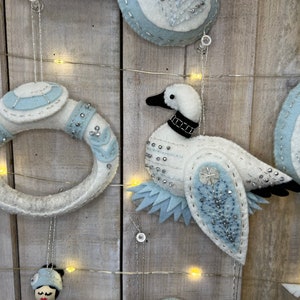 May include: A set of three felt ornaments featuring a white goose with blue and silver accents, a white ring with blue and silver accents, and a white egg with blue and silver accents.