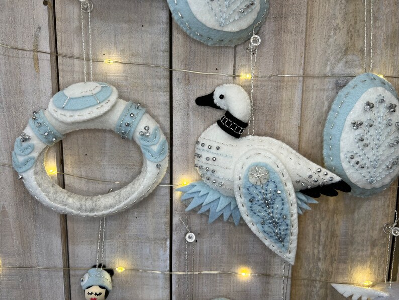 May include: A set of three felt ornaments featuring a white goose with blue and silver accents, a white ring with blue and silver accents, and a white egg with blue and silver accents.