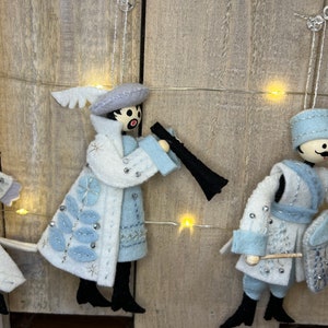 May include: Three felt Christmas decorations featuring figures in blue and white attire. The figures are playing musical instruments. The decorations are hanging from a string of fairy lights.