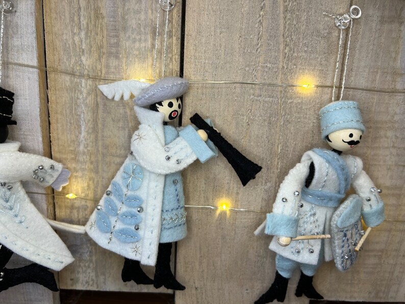 May include: Three felt Christmas decorations featuring figures in blue and white attire. The figures are playing musical instruments. The decorations are hanging from a string of fairy lights.