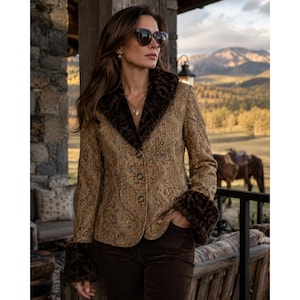 May include: A tan blazer featuring an intricate paisley pattern, a dark brown faux fur collar, and matching dark brown faux fur cuffs. The jacket is fastened with three buttons down the front and is paired with dark brown trousers and black sunglasses.