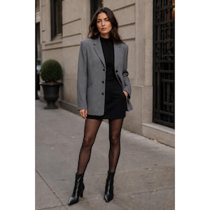 May include: A grey blazer worn over a black mock neck top and a black mini skirt. The outfit is paired with sheer black tights and black pointed-toe ankle boots. The blazer features a notched lapel, three front buttons, and two side pockets.
