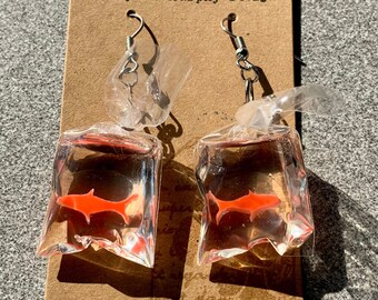 May include: A pair of dangle earrings featuring a clear plastic bag with a red fish inside. The earrings are silver and have a hook closure. The earrings are on a brown card with the text "Payton Murphy Design".