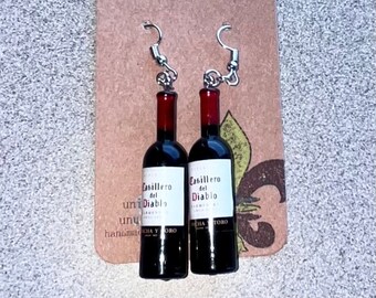 May include: A pair of earrings shaped like miniature wine bottles. The bottles are black with red tops and white labels that read "Casillero del Diablo." The earrings hang from silver-colored hooks and are displayed on a brown card with the text "Payton Murphy Design."