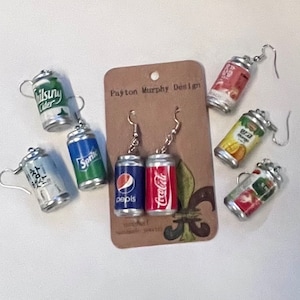 May include: A set of six pairs of dangle earrings featuring miniature soda cans. The cans are decorated with various brand logos, including Sprite, Pepsi, Coca-Cola, and more. The earrings are displayed on a brown card with the text "Payton Murphy Design".