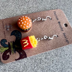 May include: A pair of dangle earrings featuring a miniature hamburger and a box of chips. The earrings are silver and have a hook fastening. The card they are displayed on reads "Payton Murphy Design" and "unique unusual handmade jewellery".