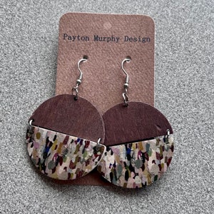 Puede incluir: Un par de pendientes de madera con la mitad superior marrón y la mitad inferior con un patrón abstracto colorido. Los pendientes están unidos a ganchos plateados. La tarjeta detrás de los pendientes dice "Payton Murphy Design".