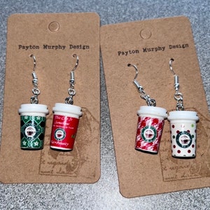 May include: Two pairs of dangle earrings featuring miniature coffee cups. One pair has a red cup with white and green designs and the text "This Cup Is Somehow Offensive Than Poverty". The other pair has a green and white cup and a black and white cup with polka dots.