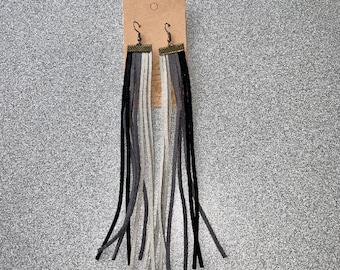 May include: A pair of long, black and gray tassel earrings with a bronze-colored top. The earrings are hanging from a brown card.