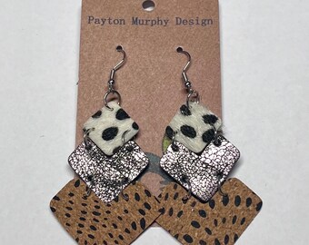 May include: A pair of dangle earrings with a layered design. The top layer is a white and black cowhide print, the middle layer is silver, and the bottom layer is a brown and black leopard print.