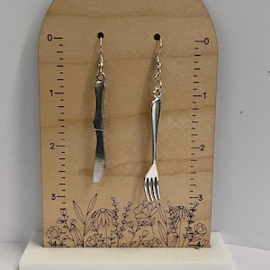 May include: A pair of silver earrings, one shaped like a knife and the other like a fork, hanging from a wooden display with a ruler and floral design.