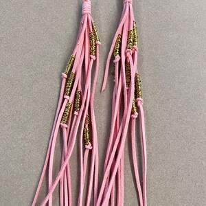 May include: A pair of pink leather tassel earrings with gold metal accents. The tassels are made of multiple strands of leather and the gold accents are small, round beads.