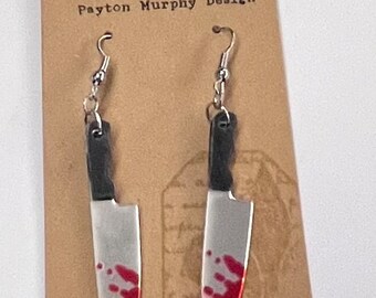 May include: A pair of silver earrings shaped like bloody kitchen knives. The earrings are on a brown card with the text "Payton Murphy Design".