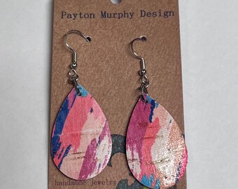 May include: A pair of teardrop-shaped cork earrings with a pink, blue, and white abstract paint design. The earrings are on a card that says "Payton Murphy Design" and "handmade jewelry".