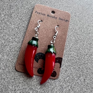 May include: Red chilli pepper dangle earrings with green tops and silver hooks. The earrings are displayed on a card with the text "Payton Murphy Design".