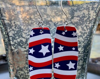 May include: A pair of patriotic earrings with a rectangular shape. The earrings feature a design of red and white stripes, blue fields, and white stars. The earrings hang from silver-colored hooks.