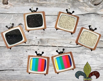 May include: Five pairs of wooden television-shaped earrings. Two pairs feature black and gold glitter, one pair features white glitter, and two pairs feature colourful stripes.