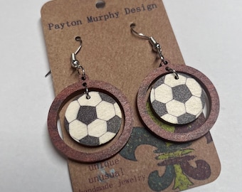 May include: A pair of brown wooden earrings with a silver hook. Each earring features a smaller circle with a black and white soccer ball design inside a larger circle.