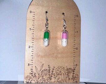May include: A pair of novelty earrings featuring a pill design. One earring has a green top and the other has a pink top, both with a white base and a smiling face. The earrings hang from silver-colored hooks.