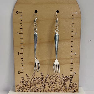 May include: A pair of silver fork earrings hanging on a wooden display with a ruler and floral design. The earrings are approximately 7.5 cm long.