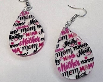 May include: A pair of teardrop-shaped earrings with a white background and pink and black lettering. The lettering includes the words "Mom", "Mother", "Mommy", "Mama", and "Madre".