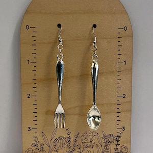May include: A pair of silver earrings, one shaped like a fork and the other like a spoon, hanging from a wooden display with a floral design and a ruler.