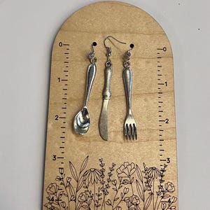 May include: Silver-coloured spoon, knife, and fork earrings. The earrings are displayed on a wooden board with a ruler and floral design. The earrings are approximately 5 cm in length.