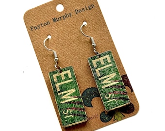 May include: A pair of wooden earrings featuring a green street sign design with the text "ELM St." in white. The earrings hang from silver-colored hooks and are displayed on a brown card with the text "Payton Murphy Design".