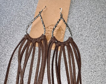 May include: Brown suede fringe earrings with silver teardrop-shaped hoops. The earrings have a bohemian style.