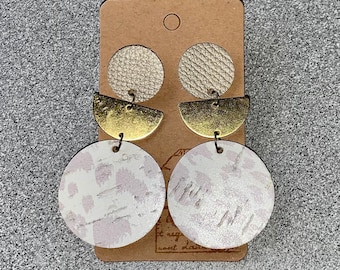 May include: A pair of gold and white leather earrings with a geometric design. The earrings are made of two circles, one smaller than the other, with a half-circle shape in between. The smaller circle is white with a pink and white pattern, and the larger circle is white with a gold and white pattern. The half-circle is gold.