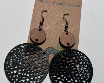 May include: A pair of black leather earrings with a circular cutout design. The earrings have a brown wooden accent at the top and a bronze hook.