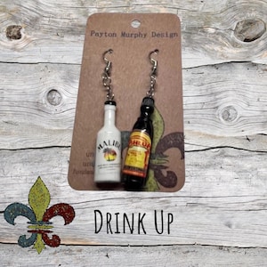 Könnte beinhalten: Ein Paar silberne Ohrhänger mit Miniatur-Likörflaschen. Die Flaschen sind weiß und schwarz mit den Etiketten "Malibu" und "Kahlua". Die Karte hinter den Ohrringen zeigt "Payton Murphy Design" und "Drink Up".